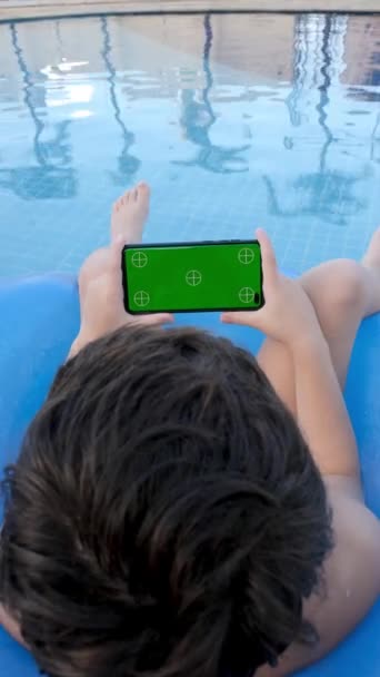 9岁的孩子坐在浮标上 水平观看装有绿色屏幕的智能手机 — 图库视频影像
