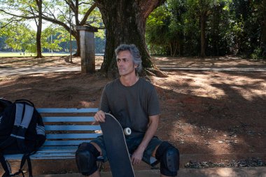 Brazilian skateboarder over 50 years old relaxing on a bench in a square_1. clipart
