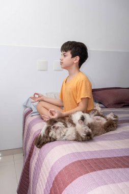 11 yaşındaki Brezilyalı çocuk yatağında oturmuş yoga yapıyor ve gözleri köpeğinin yanında açık olarak duruyor..