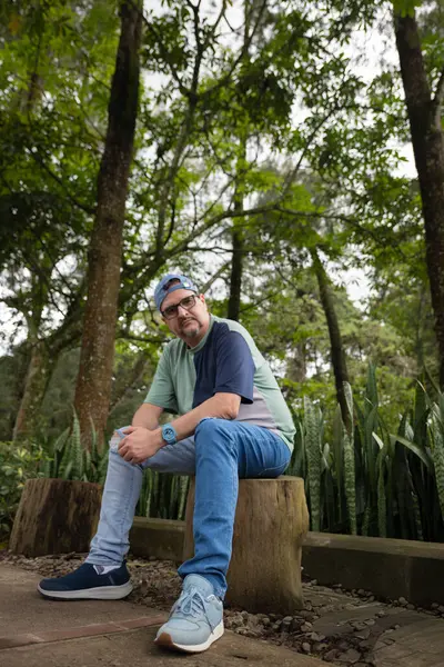 Mavi sakallı ve gözlüklü bir gencin portresi, günlük kıyafetler giymiş ormanda oturuyor ve dinleniyor..