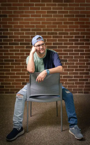 Gözlüklü ve mavi tişörtlü genç yakışıklı adam sandalyede oturuyor.