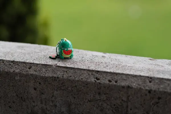 Küçük yeşil oyuncak bir dinozor anahtarlığının yakın plan görüntüsü. Bir dilim karpuz yiyor. Bulanık yeşil arka planlı, taş bir yüzeye yerleştiriliyor. Çocukluk kavramı, eğlence, yaz neşesi ve eğlenceli hayal gücü..