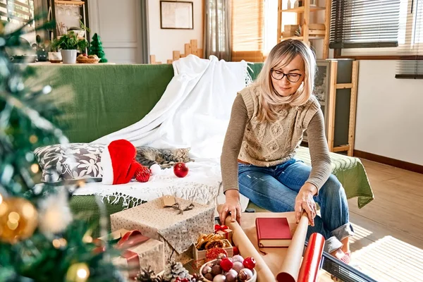 Noel hediyelerini paketliyorum. Sarışın bir kadın geri dönüşümlü kartla kitap sarıp Noel ağacının yanında kurutulmuş portakallar ve köknar dallarıyla süslüyor. Kış bayramı kutlaması.