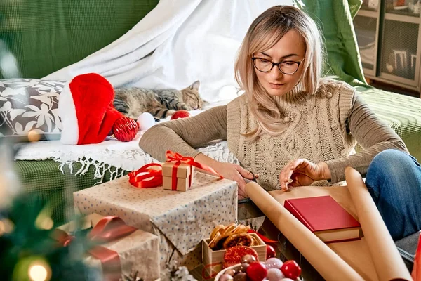 stock image Wrapping christmas gifts. Blond woman wrapping a book as present in recycled card and decorate it with dried oranges and fir branches near the Christmas tree. Winter holiday celebration.