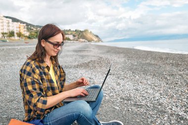 Ekose gömlekli, laptoplu bir kadın açık havada çalışırken deniz kenarında kış kumsalında oturuyor. Modern yaşam tarzı, bağlantı, blog yazma, iş dünyası, serbest çalışma.