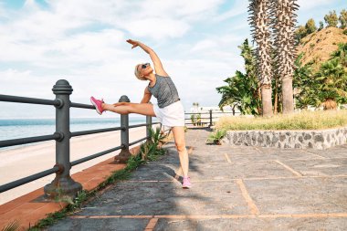 Young beautiful sportive woman training over seaside promenade, stretching legs before jogging. Fitness, sport and healthy lifestyle concept.