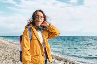 Smiling woman in yellow jacket with backpack and coffee mug portable walking along sea coast and enjoying the sunny day. Concept of hiking and activity in the low season.