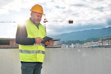 Portrait of middle aged bearded supervisor in hardhat and safety vest with tablet on building site. Structural engineer or architect monitors the progress of the work on construction site.