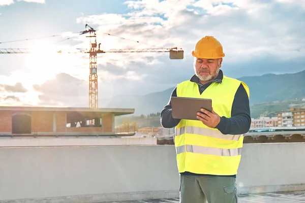 Middle aged bearded supervisor in hardhat and safety vest making notes in tablet on building site. Structural engineer or architect monitors progress of work on construction site.