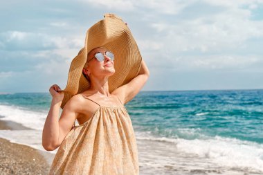 Pretty blond woman in huge beach hat reaches out to the sun, enjoying sunny windy day near blue sea. Travel concept. Pastel muted color.