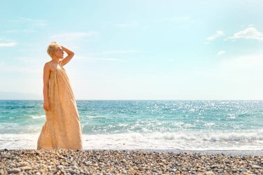 Pretty blond woman walking on pebble beach enjoying sunny windy day near blue sea. Travel concept. Pastel muted color.