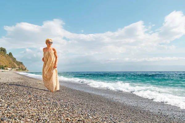 stock image Pretty blond woman walking on pebble beach enjoying sunny windy day near blue sea. Travel concept. Pastel muted color.