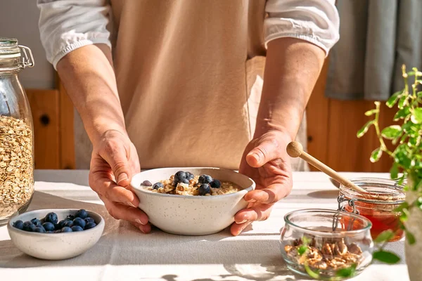 Woman preparing healthy dieting vegan nutritious breakfast. Female hands holding bowl of oatmeal porridge with blueberries, walnuts and honey.