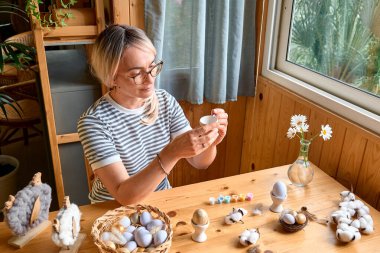 Sarışın kadın evde paskalya süsü hazırlıyor, renkli paskalya yumurtaları ve renkli yumurta fincanları boyuyor..