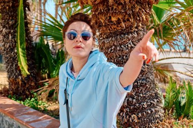 Güneş gözlüğü takan ciddi bir kadın sanki deniz kıyısında görünmeyen bir şeye dokunuyormuş gibi uzanıyor. Huzurlu doğada özgürlüğün tadını çıkaran gezgin. Ruh sağlığı, sağlık, macera yolculuğu ve sağlıklı yaşam tarzı.