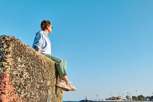 Young woman sitting on big stone in seaside with blue sky in background and enjoying freedom. Female traveler relaxing in serene nature. Mental health, wellbeing, trip adventure and healthy lifestyle.