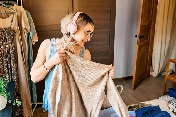 Changing seasonal clothes. Blond woman in headphones listening music, dancing and singing while making order of clothes arranging garments in the box in the bedroom with hanger rack and wardrobe.