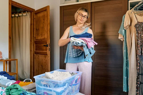 Changing seasonal clothes. Blond woman making order of clothes arranging garments in the box in the bedroom with hanger rack and wardrobe.