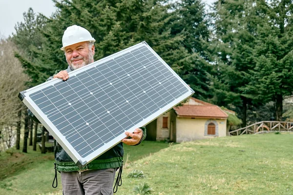 Middle aged bearded technician with voltaic solar panel near house in rural zone. Stand-alone photovoltaic solar panel system. Renewable ecological cheap green energy production. Energy independence.