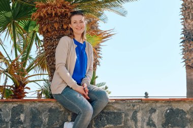 Kameraya bakarak gülümseyen sarışın kadının portresi deniz kenarında palmiye ağacının yanında özgürlüğün tadını çıkarıyor. Dişi gezgin sakin doğada dinleniyor. Akıl sağlığı, refah, yolculuk macerası ve sağlıklı yaşam tarzı.