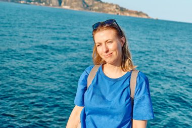 Kameraya bakıp deniz kenarında özgürlüğün tadını çıkaran gülümseyen sarışın kadının portresi. Dişi gezgin sakin doğada dinleniyor. Akıl sağlığı, refah, yolculuk macerası ve sağlıklı yaşam tarzı..