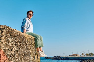 Sahilde büyük bir taşın üzerinde oturan genç bir kadın. Arka planda mavi gökyüzü ve özgürlüğün tadını çıkarıyor. Dişi gezgin sakin doğada dinleniyor. Akıl sağlığı, refah, yolculuk macerası ve sağlıklı yaşam tarzı..