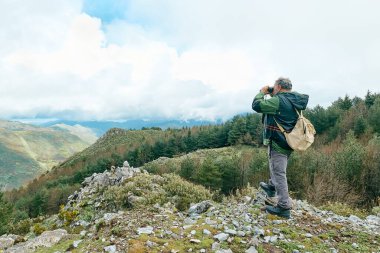 Dağ ormanlarında yürüyüş yapan gezgin adam, kayanın üzerinde durup dürbünle panoramik dağ vadisine bakıyor..