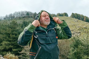 Dağ ormanlarında yürüyüş yapan, dağ vadisinde rüzgardan korunmak için kapüşonlu ceket giyen olgun sakallı bir gezgin..