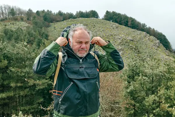 stock image Mature bearded traveler man hiking in mountain forest, wearing jacket with hood to protect himself from the wind in the mountain valley.