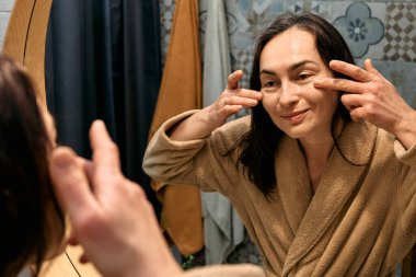 Bornozlu orta yaşlı bir kadın banyoda aynaya bakıyor ve yüzüne dokunuyor, cildine yaşlanma önleyici ilaçlar sürüyor ve gülümsüyor..