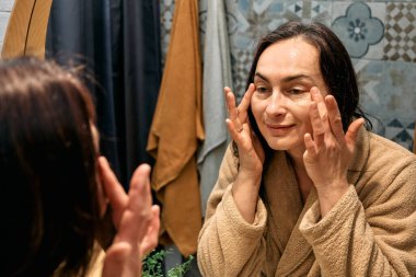 Bornozlu orta yaşlı bir kadın banyoda aynaya bakıyor ve yüzüne dokunuyor, cildine yaşlanma önleyici ilaçlar sürüyor ve gülümsüyor..