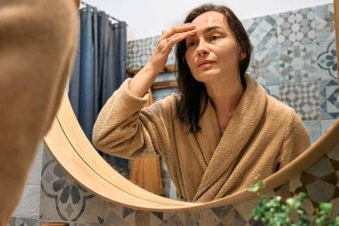 Bornozlu orta yaşlı bir kadın banyoda aynaya bakıyor ve yüzüne dokunuyor, cildine yaşlanma önleyici ilaçlar sürüyor ve gülümsüyor..