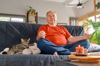Kilolu bir adam evde yoga ve meditasyon yapıyor. Lotus 'ta oturuyor, kanepeye oturuyor, gözleri kapalı rahatlıyor. Dikkatli meditasyon konsepti. İyi olmak..
