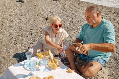 Yaz pikniği. Orta yaşlı çift sahilde taze meyve, şarap ve aperatiflerle piknik yapıyor. Sakallı adam, sarışın kadın için şarap kadehine şarap dolduruyor..