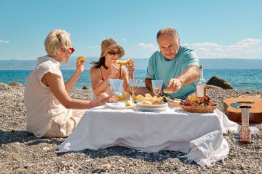 Yaz pikniği. Bir grup orta yaşlı arkadaş sahilde taze meyve, şarap ve aperatiflerle piknik yapıyor, şarap içiyor, konuşuyor, eğleniyor, yemek yiyor..