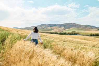 Arkadaki kadın altın buğday tarlasında yürüyor sıcak yaz güneşinde ve mavi gökyüzünde arkasında dağ tepeleri olan beyaz bulutlarla.