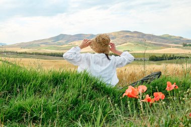 Hasır şapkalı kadın, haşhaş çayırında oturuyor ve sıcak yaz güneşinde altın buğday tarlasına hayran. Ve mavi gökyüzü, arka planda dağ tepe manzaralı beyaz bulutlar..