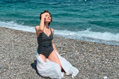 Mayo giymiş güzel seksi bir kadın ve mavi deniz kenarında güneşli bir havanın tadını çıkaran beyaz bir pareo. Hayattan zevk alan mutlu bir kadın. Plaj yazı, seyahat konsepti.