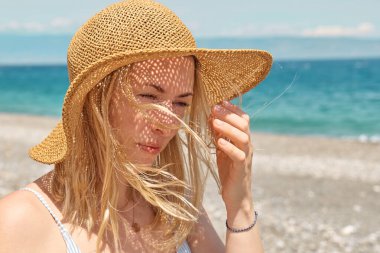 Plaj şapkalı güzel sarışın kadının portresi mavi deniz kenarında güneşli ve rüzgarlı bir günün tadını çıkarıyor. Hayattan zevk alan mutlu bir kadın. Plaj yazı, seyahat konsepti.