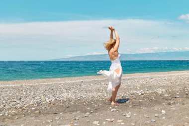 Mayo giymiş güzel sarışın bir kadın ve mavi deniz kenarında güneşli bir günün tadını çıkaran beyaz bir pareo. Hayattan zevk alan mutlu bir kadın. Plaj yazı, seyahat konsepti.