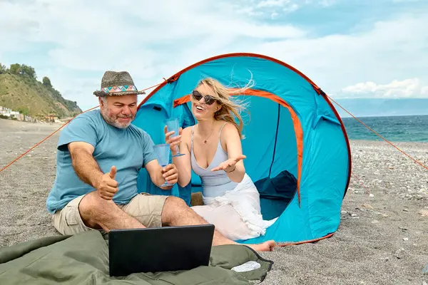 Couple having fun, drinking wine and talking with friends in video call in laptop in picnic near tent on seaside. Vacation summer on the beach in outdoors. Travel, vacation and friendship concept.