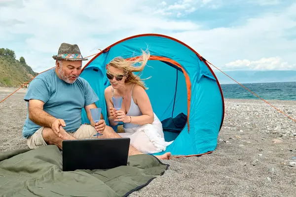 Couple having fun, drinking wine and cheer the match video on laptop during picnic near tent by the sea. Summer beach vacation outdoors.