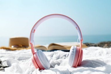 Yaz tatilinde Audiobook ya da podcast. Kulaklıkları açık kitap, güneş gözlüğü ve arka planda deniz kenarı olan şapkaların yanına kapat..