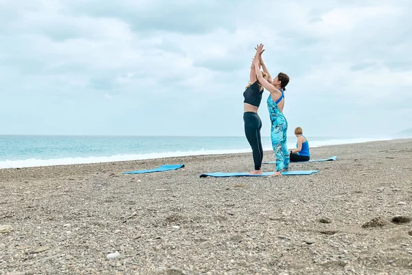 Summer pilates training. Group of women with fitness instructor practicing yoga exercise at the beach near the water. Yoga retreat workout, body and mind wellness at the sea.