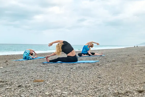 Summer pilates training. Group of women practicing yoga exercise at the beach near the water. Yoga retreat workout, body and mind wellness at the sea.