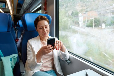 Gülümseyen iş kadını serbest blog yazarı trende cep telefonu kullanıyor. Trenle seyahat eden güzel bir kadın pencere kenarında oturan akıllı telefonu iş için ya da sosyal medya için kullanıyor..