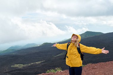 Avrupa 'nın en yüksek volkanı olan Etna' da yürüyüş. Sarı yağmurluklu genç bir kadın dağın zirvesinde kollarını kaldırıp Etna Dağı 'nın panoramik manzarasına bakıyor..