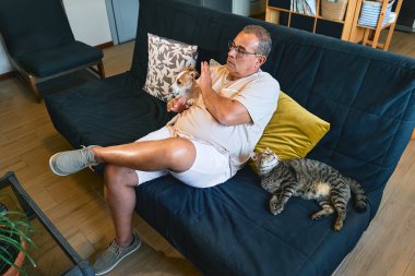 Orta yaşlı bir adam kanepede kedi köpek çiftliğinde evcil hayvanlara sarılıp televizyon seyrederek vakit geçiriyor. Jack Russell köpek yavrusu insan yüzünü yalıyor.