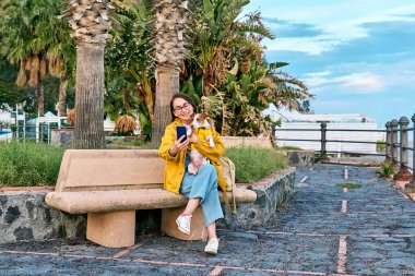 Sarı yağmurluk giymiş ve bir bahar ya da sonbahar günü deniz kenarında yürürken akıllı telefonuyla selfie çeken bir kadın..