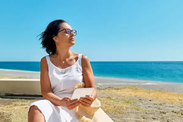 Mature woman wearing using digital tablet at the beach. Middle aged female using digital device for shopping online, banking, social media or reading ebook outdoors.